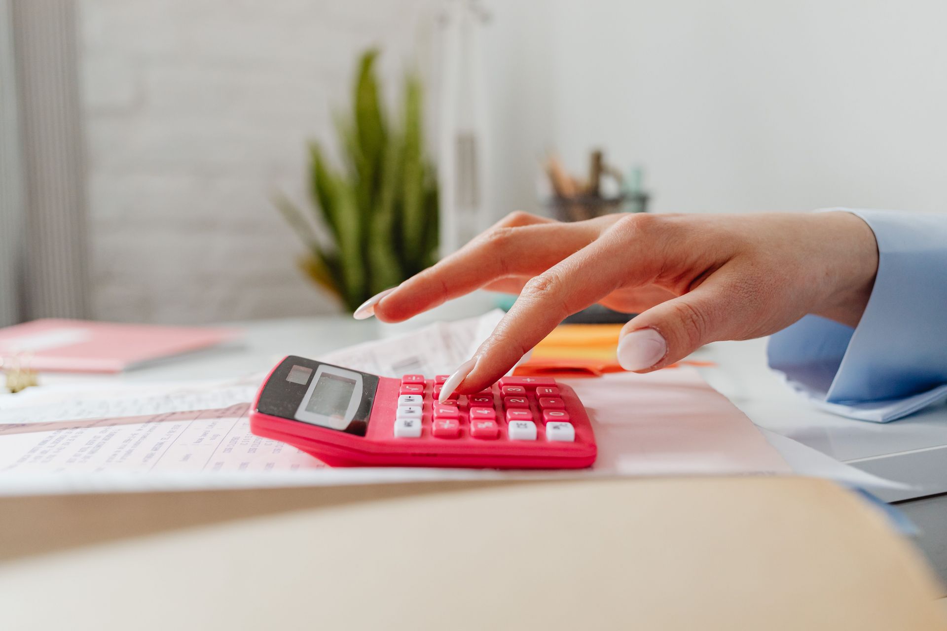 A close-up image of a person's hand using a red calculator on a desk with paperwork.